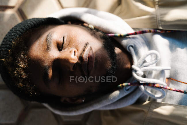 Top view of calm young bearded African American male in trendy outfit lying on ground with closed eyes on sunny day — Stock Photo