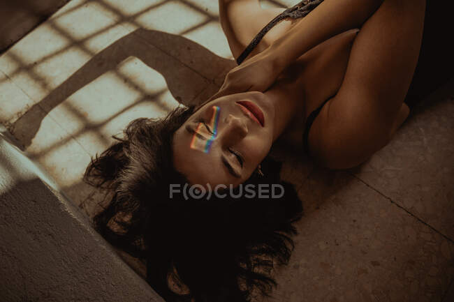 High angle of serene female with rainbow light on face lying on floor with closed eyes in room with sunlight — Stock Photo