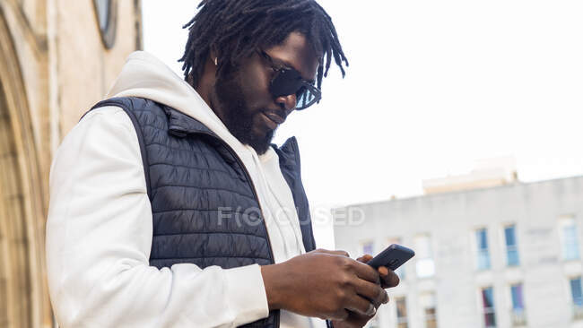 From below trendy African American male in sunglasses browsing on mobile phone while standing building in town — Stock Photo