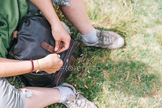 High angle crop anonymous female in casual wear sitting on grass and closing leather backpack while camping in sunny nature — Stock Photo