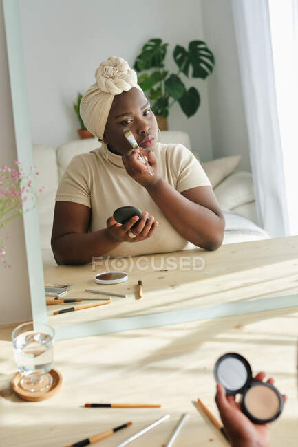 Elegante jovem fêmea africana robusta em turbante tradicional aplicando fundação no rosto enquanto está perto do espelho na luz moderna quarto — Fotografia de Stock
