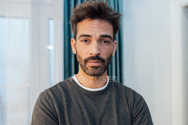 Concentrated young bearded ethnic male in casual clothes standing in modern apartment and looking at camera — Stock Photo