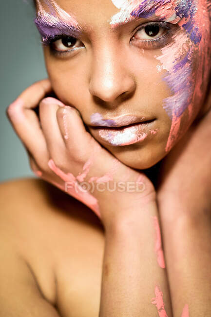 Modelo feminino étnico criativo com rosto manchado com tinta rosa e branca tocando bochechas e olhando para a câmera em fundo cinza no estúdio — Fotografia de Stock