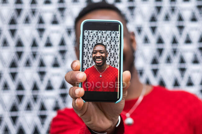 Macho étnico em roupas brilhantes cobrindo rosto com celular enquanto demonstra foto na tela durante o dia — Fotografia de Stock