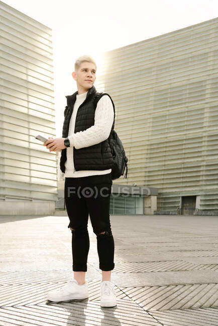 Side view of young stylish guy in trendy knitted sweater browsing on her smartphone while waiting for appointment on city street near contemporary buildings on sunny day — Stock Photo