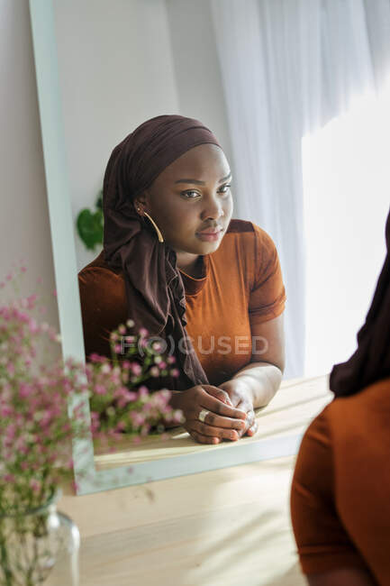 Confident young stylish African lady in traditional hijab leaning on cosmetic table and looking in mirror in sunlight at home — Stock Photo