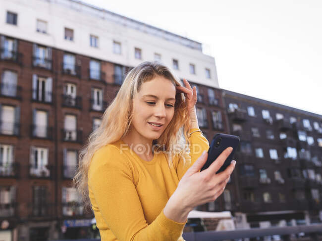 Mujer joven positiva con el pelo rubio ondulado tocando el pelo y sonriendo mientras tiene videollamada en el teléfono móvil en la plaza de la ciudad - foto de stock