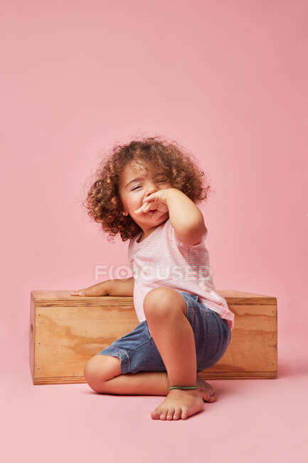 Charmant enfant pieds nus joyeux en t-shirt et short en denim avec des cheveux bouclés assis sur le sol en jouant sur une plate-forme en bois — Photo de stock