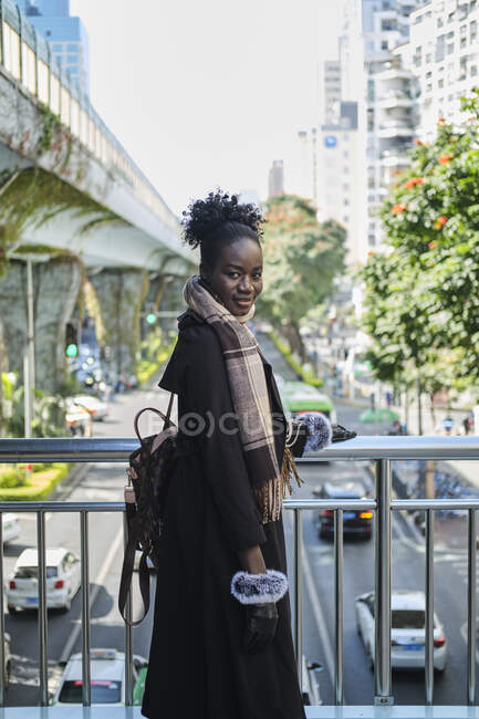Seitenansicht einer jungen lächelnden schwarzen Frau in stylischer Kleidung mit Rucksack, die von einer Brücke über der Stadtstraße in die Kamera blickt — Stockfoto