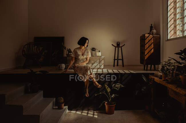Нежная женщина в платье сидит в комнате с различными растениями в горшках дома — стоковое фото