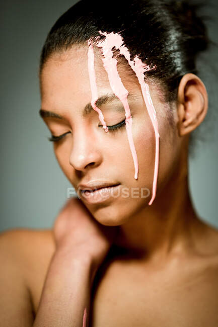 Creative ethnic female model with pink paint dripping on her face with eyes closed on gray background in studio — Stock Photo