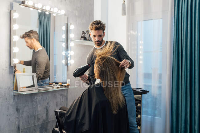 Ethnique homme coiffeur séchage des cheveux de la cliente avec les yeux fermés dans le studio de beauté moderne — Photo de stock