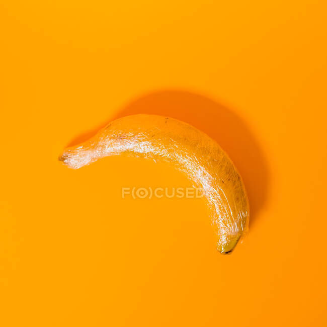 Зверху смачний стиглий банан, покритий прозорою пластиковою обгорткою, що представляє концепцію промислового сільського господарства на яскраво-жовтому тлі — стокове фото