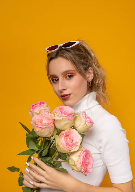 Молодая красивая женщина в белом наряде и модных солнцезащитных очках, держа нежные розовые розы, стоя на желтом фоне в фотостудии — стоковое фото