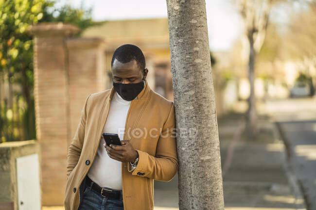 Неузнаваемый вдумчивый молодой чернокожий мужчина в модном наряде и маске для лица, опирающийся на дерево на городской улице во время телефонного разговора — стоковое фото