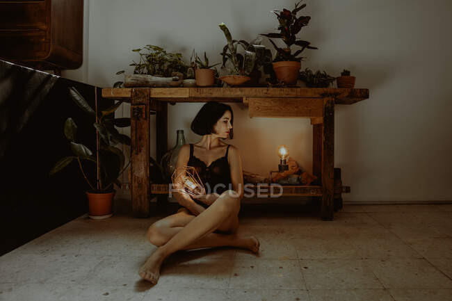 Tranquille pieds nus femme assise sur le sol avec lanterne lumineuse dans la chambre avec des plantes en pot et regardant loin — Photo de stock