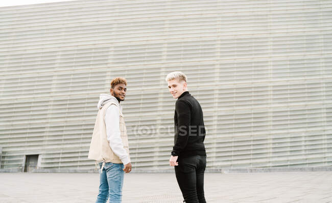 Side view of happy young multiracial male friends in trendy outfits standing on city street near modern building and looking over shoulder — Stock Photo