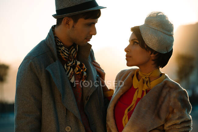 Hombre de la cosecha en ropa de moda cerca de la pareja femenina en sombrero vintage mirándose por la noche - foto de stock