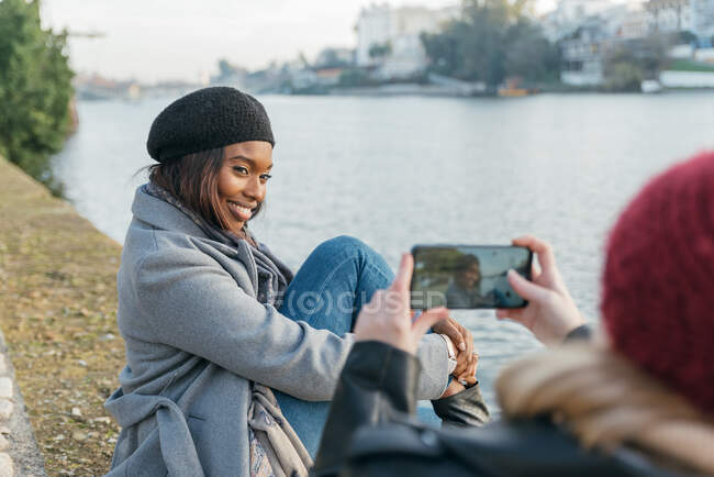 Mujer irreconocible tomando una foto de una amiga negra en un teléfono inteligente mientras se relaja en el terraplén en la ciudad - foto de stock