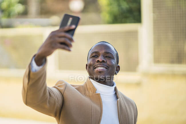 Feliz joven afroamericano en traje de moda en la calle y haciendo un autorretrato con su teléfono inteligente en la calle en el soleado día de verano - foto de stock