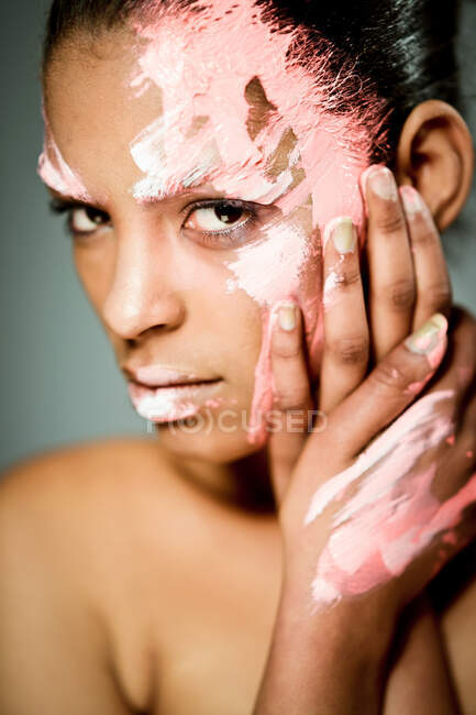 Modèle féminin ethnique créatif avec visage enduit de peinture rose et blanche touchant les joues et regardant la caméra sur fond gris en studio — Photo de stock