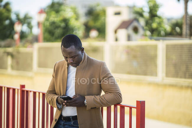 Concentrated young African American male in trendy outfit leaning on fence on street and browsing mobile phone on sunny summer day — Stock Photo