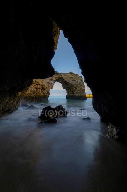 Através de uma vista pitoresca das formações rochosas arqueadas no litoral oceânico sob céu azul na Praia da Abandeira, Algarve Portugal — Fotografia de Stock