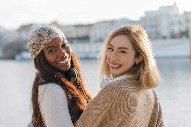 Guardando la fotocamera due amici femminili multirazziali in cuffia godendo insieme su argine nella giornata di sole — Foto stock