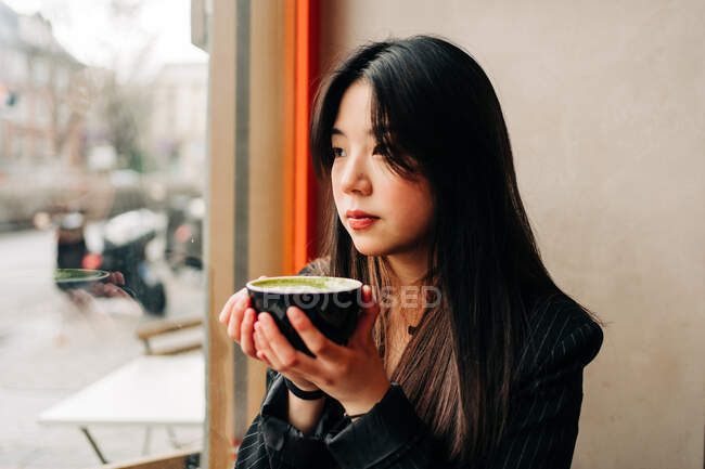Morena de cabelos compridos Mulher asiática tomando um café em uma cafeteria enquanto olha um celular pela janela — Fotografia de Stock