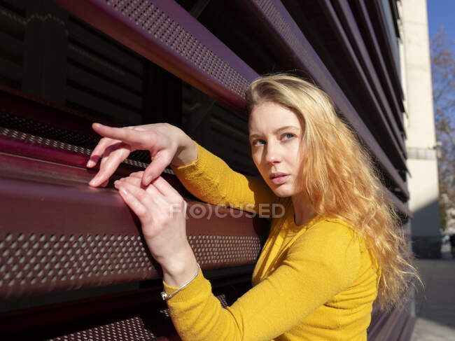 Vista lateral de la joven milenaria pensativa en ropa casual con el pelo ondulado largo y rubio de pie cerca de la valla de metal rojo - foto de stock