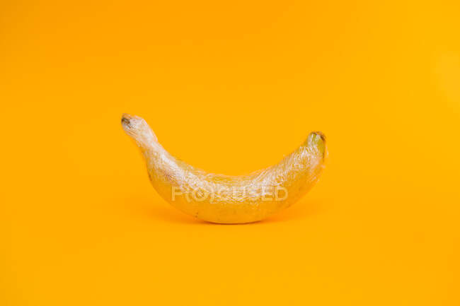 Köstliche reife Banane mit transparenter Plastikfolie bedeckt, die das Konzept der industriellen Landwirtschaft auf leuchtend gelbem Hintergrund darstellt — Stockfoto