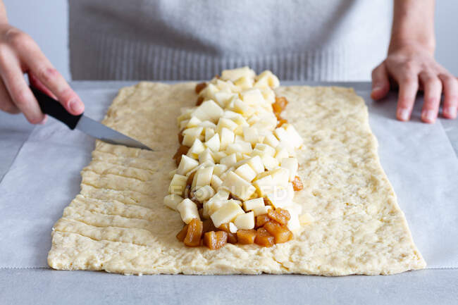 Анонимная женщина-повар урожая режет сырое тесто вкусными свежими и приготовленными яблочными ломтиками, готовя плетеную выпечку на бумаге — стоковое фото
