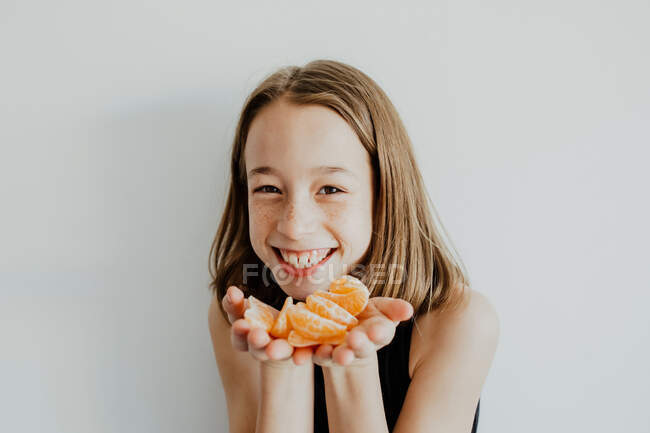 Обрізати позитивну дівчину з веснянками посміхаючись і дивлячись на камеру, демонструючи скибочки свіжого здорового мандарина — стокове фото