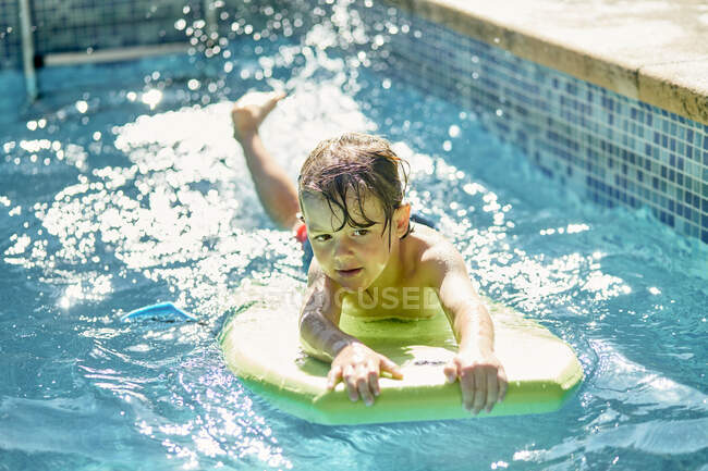 Высокий угол внимательный милый мальчик лежит на тренировочной доске во время плавания в открытом бассейне в солнечный день — стоковое фото
