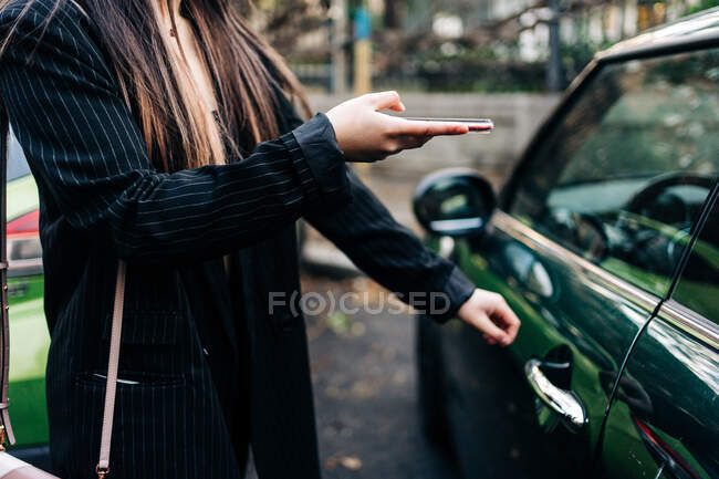 Неузнаваемая длинноволосая женщина, открывающая машину с помощью мобильного телефона — стоковое фото