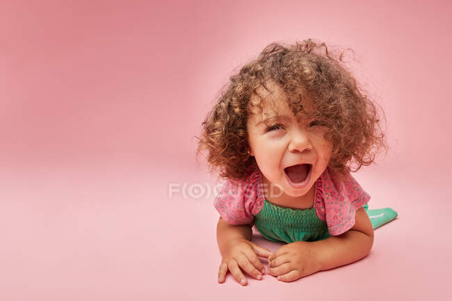 Entzückendes Kleinkind im Kleid mit lockigem Haar, das einen Wutanfall hat und wegschaut, während es auf dem Boden sitzt — Stockfoto