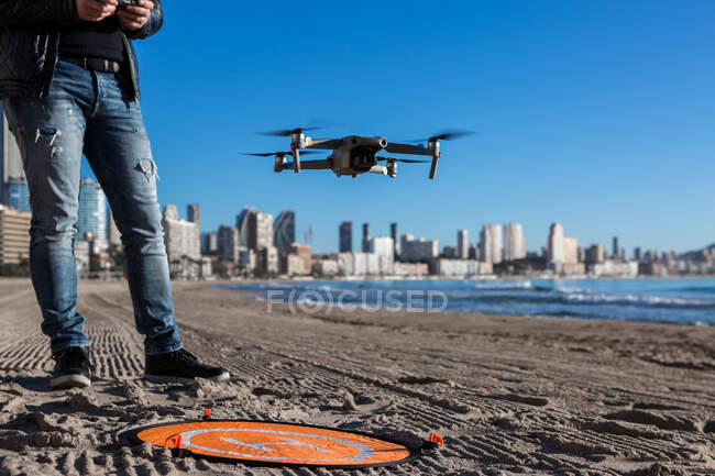 Desde arriba de macho no reconocible recortado con mando a distancia de pie en la playa con dron colocado en la plataforma de aterrizaje - foto de stock