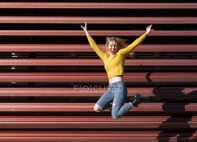 Cuerpo completo de alegre joven milenaria en traje casual saltando alto con los brazos levantados y sonriendo cerca de valla de metal en la calle - foto de stock