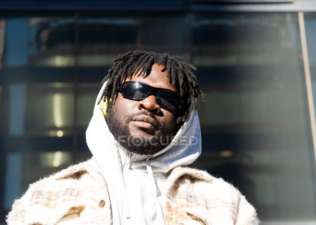 Young contemplative bearded African American male in trendy sunglasses with dreadlocks looking at camera in city — Stock Photo