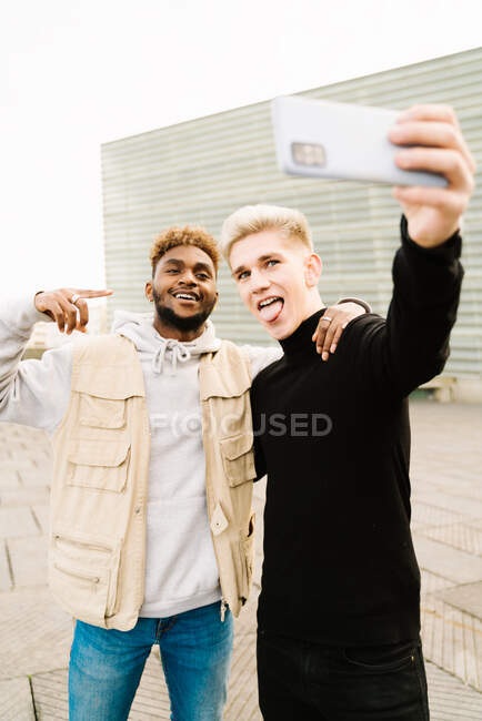 Щасливий молодий афроамериканець у модному одязі посміхається, коли робить автопортрет на вулиці з рукою на плечі друга-чоловіка і язиком — стокове фото