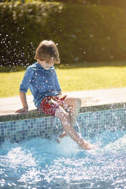 Bambino irriconoscibile seduto sul bordo della piscina all'aperto e spruzzi d'acqua con le gambe nella soleggiata giornata estiva — Foto stock