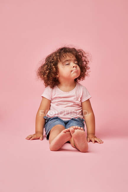 Cute toddler girl with curly hair in casual clothes having fun making faces while sitting on the floor with eyes closed on pink background — Stock Photo