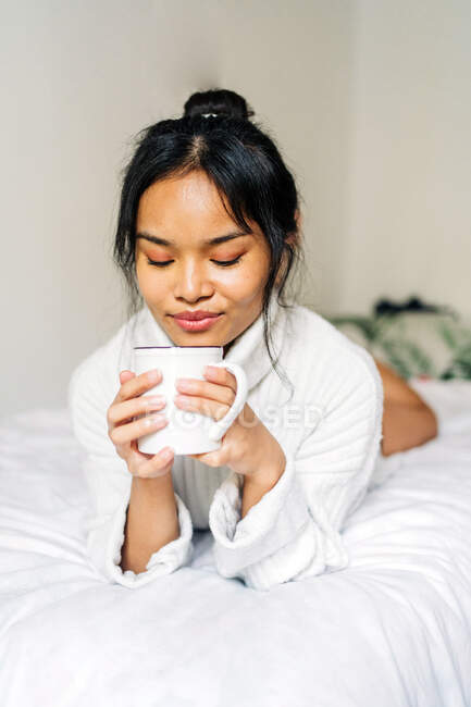 Cabello largo morena asiática mujer acostada en la cama en casa con una taza de café - foto de stock