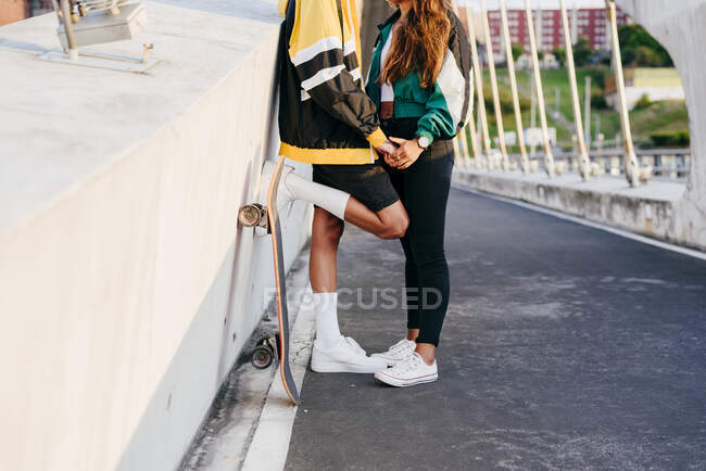 Cultivado pareja irreconocible con traje urbano y monopatín acostado en una pared en la calle - foto de stock