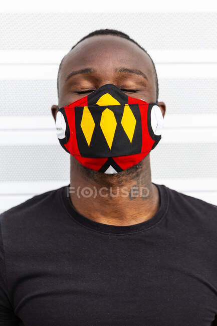 Anonymous young African American male with closed eyes in bright mask with ornament during coronavirus period on light background — Stock Photo