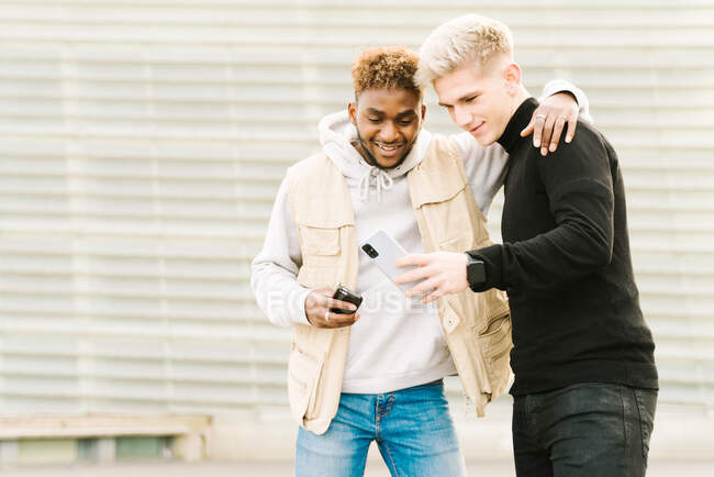 Щасливий афроамериканець у модному одязі посміхається, стоячи на вулиці з рукою на плечі друга-чоловіка, який показує відео на смартфоні. — стокове фото