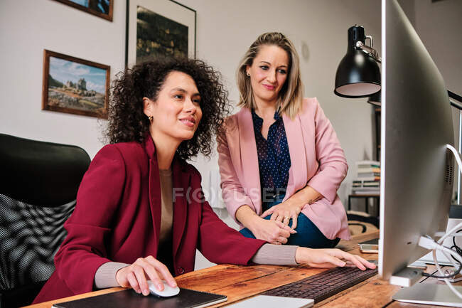 Весёлые многорасовые коллеги-женщины в формальном наряде делятся настольным компьютером, взаимодействуя в современном рабочем пространстве — стоковое фото