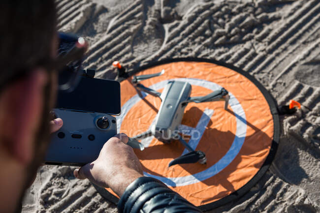 Desde arriba de macho irreconocible con mando a distancia de pie en la playa con dron colocado en la plataforma de aterrizaje - foto de stock