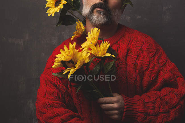 Unrecognizable creative mature male in knitted sweater covering face with bright sunflowers against black background — Stock Photo