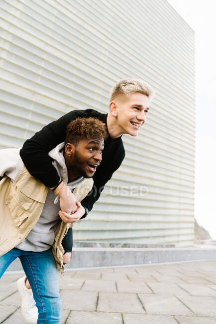 De baixo de menino negro jovem alegre dando passeio de piggyback para amigo alegre enquanto passam o tempo juntos no parque da cidade — Fotografia de Stock